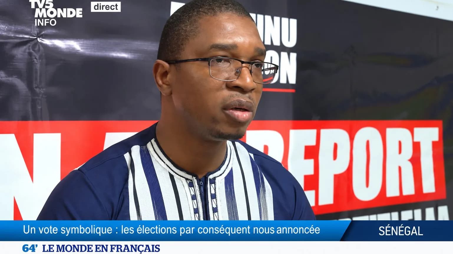 Jean Aimé interrogé sur le plateau de TV5MONDE, devant un fond « Report » barré de rouge